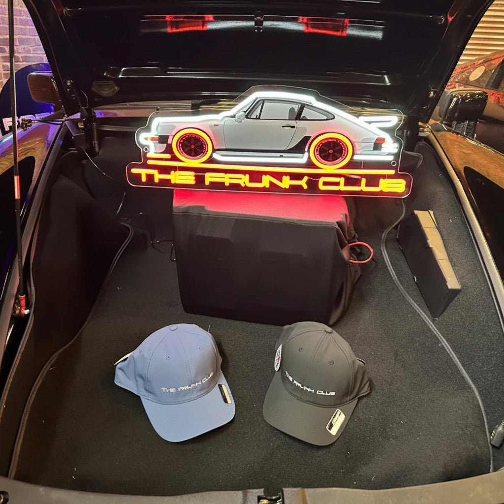 The Frunk Club Pit Lane Cap & Patch bundle featuring two customizable baseball caps in navy and grey, displayed in a car trunk with neon car sign.
